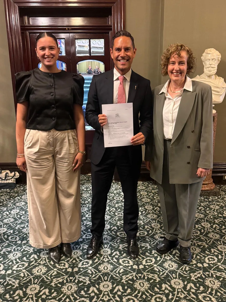 Aria Kapaniris on the left, Alex Greenwich in the middle and Teresa
                      Russell on the right, standing together inside NSW Parliament. Alex Greenwich
                      holds a printed copy of the Bill.